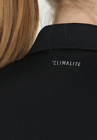 Černé polo tričko s hladkým povrchem a plochým límcem. Na zadní straně se nachází bílé logo "CLIMALITE".