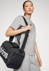 Μαύρη τσάντα duffel με λευκό λογότυπο adidas, δίχτυ ώμου και πλέγμα. Το μοντέλο φοράει ένα γκρι μπλουζάκι με τρεις μαύρες ρίγες.
