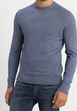 Mand iført en tætsiddende, langærmet blå rundhalsstrik med et lille logo på brystet, kombineret med mørkeblå jeans.