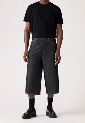 Persoon die een zwart T-shirt met korte mouwen draagt, donkere wijde cropped broek, zwarte sokken en zwarte leren loafers, staand op een lichte achtergrond.