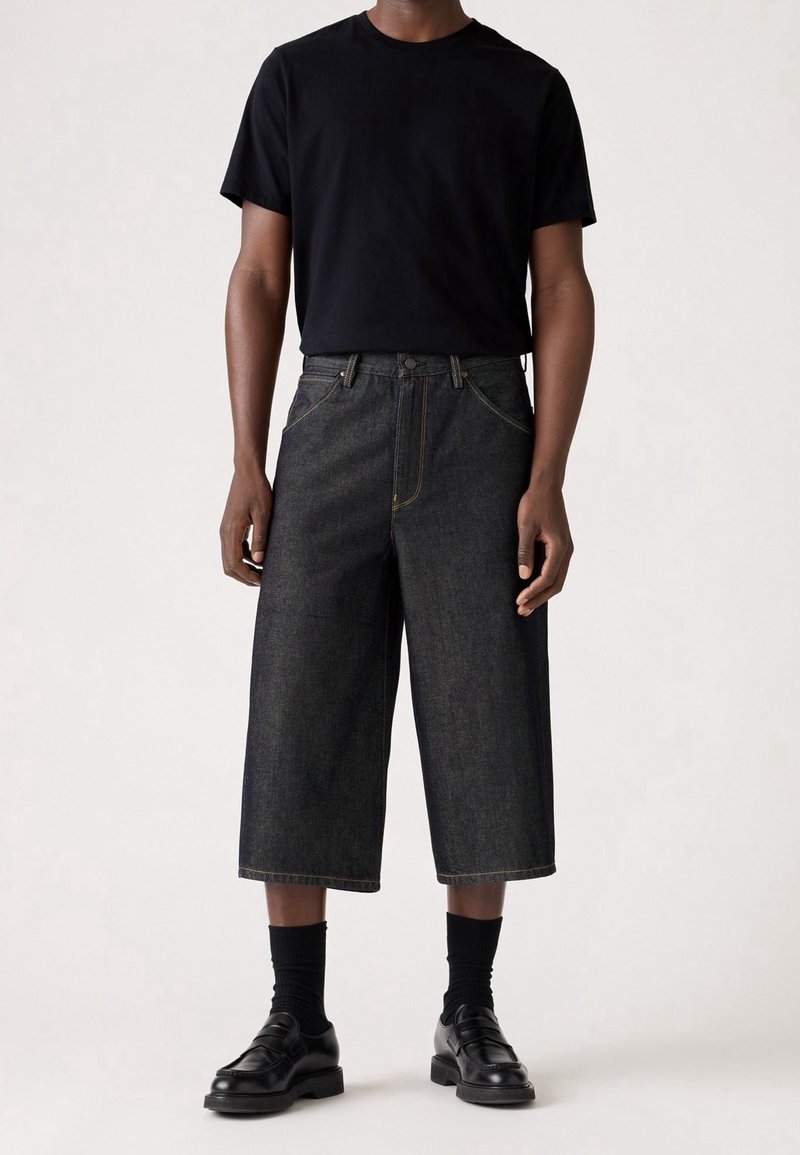 Personne portant un t-shirt noir à manches courtes, un pantalon large court sombre, des chaussettes noires et des mocassins en cuir noir, debout sur un fond clair.