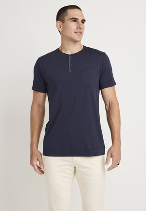Navyblaues kurzärmliges Henley-Shirt aus Baumwolle mit einer durchgeknöpften Knopfleiste und einer Brusttasche, kombiniert mit hellen Hosen.