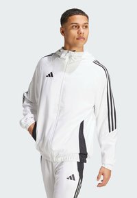 Coupe-vent blanc zippé avec des panneaux latéraux noirs et trois bandes noires sur les manches. Dispose d'une capuche ajustable et de poignets élastiques.