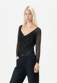 Γυναίκα με μακριά ξανθά μαλλιά, φορώντας ένα μαύρο διάφανο μπλουζάκι με μακριά μανίκια και σκούρα ψηλόμεσα τζιν, στυλ αναμένο με το ένα χέρι στην τσέπη.