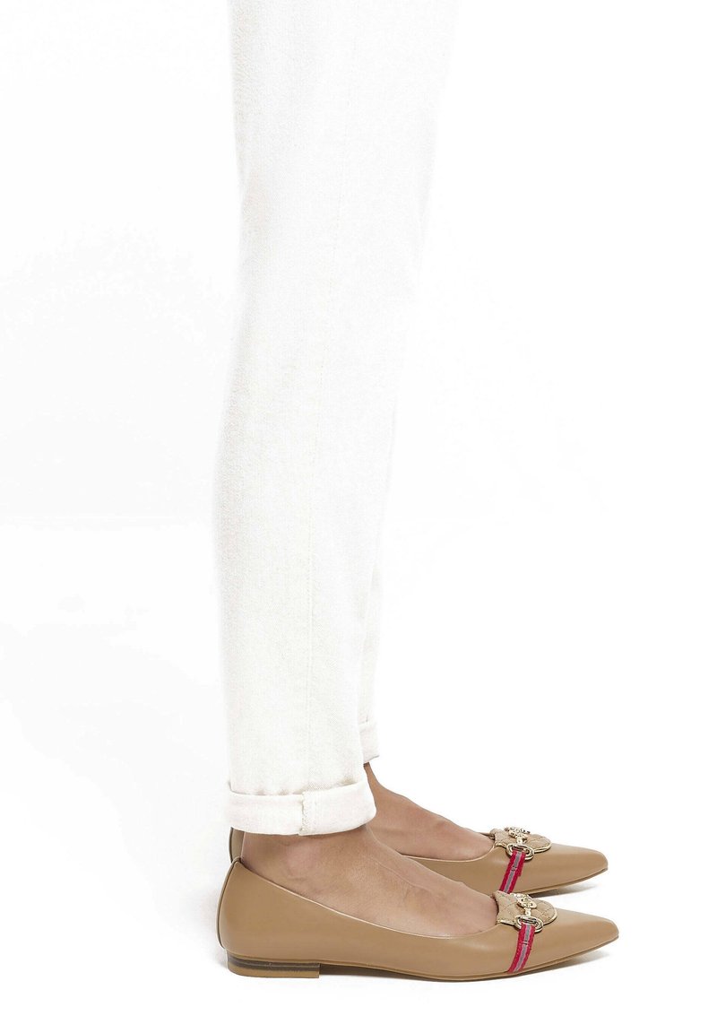 Pieds portant des ballerines beige à bout pointu avec des ornements rouges et dorés, associées à un pantalon blanc retroussé sur un fond blanc.