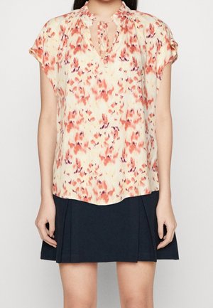Kvinde iført en kortærmet bluse med blomsterprint og bindebånd i halsen, kombineret med et sort plisseret skørt.