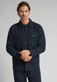 Chaqueta azul marino de botón con cuello. Presenta dos bolsillos delanteros con solapa y mangas largas, confeccionada con una tela texturizada.