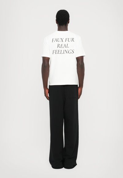 T-shirt branca com texto preto nas costas a dizer "FAUX FUR REAL FEELINGS", combinado com calças largas pretas.