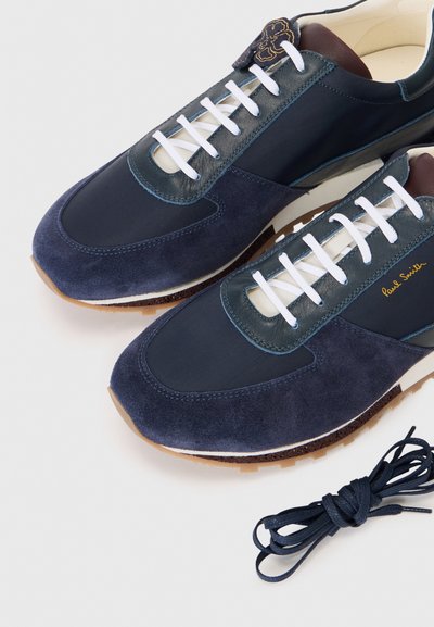 Blue suede and textile sneakers feature a padded tongue, white laces, and a brown leather accent, with a rubber sole and gold branding.