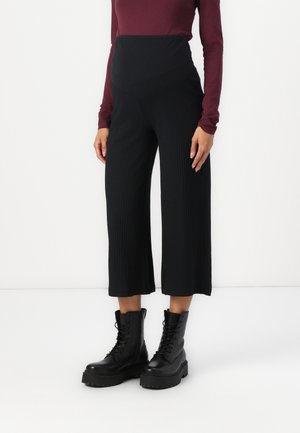 Persona con pantalones de pierna ancha negros de talle alto, botas negras estilo militar con cordones y una camiseta de manga larga color granate sobre un fondo blanco.