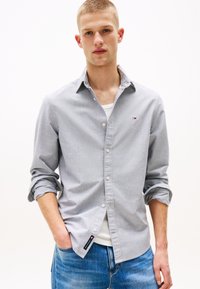 Bărbat tânăr purtând o cămașă gri deschis cu nasturi peste un tricou alb, cu mânecile suflecate și blugi albaștri, cu mâinile în buzunare.