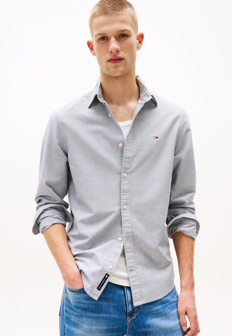 Bărbat tânăr purtând o cămașă gri deschis cu nasturi peste un tricou alb, cu mânecile suflecate și blugi albaștri, cu mâinile în buzunare.