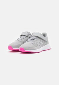 Une paire de chaussures de sport grises avec des semelles roses, à lacets à l'avant et avec une sangle à boucle, présentée sur fond blanc.