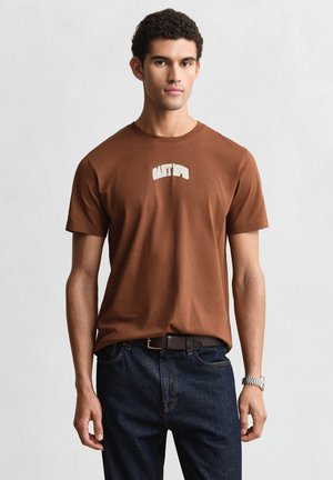 Junger Mann, der ein braunes GANT 1949 T-Shirt, dunkle Jeans mit Gürtel und eine silberne Uhr trägt, steht vor einem schlichten hellen Hintergrund.