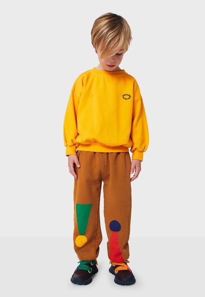 Sudadera amarilla con logo, pantalones marrones con parches geométricos coloridos y zapatillas negras con cordones multicolores.