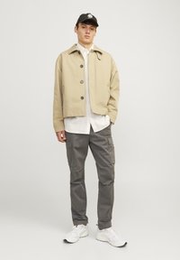 Beige kort jacka med knappstängning, lager-på-lager över en vit randig skjorta. Grå cargobyxor och vita sneakers fulländar outfiten.