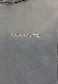 Tissu gris avec texte brodé en blanc "DROPSIZE" centré sur la surface, avec des détails de coutures visibles.