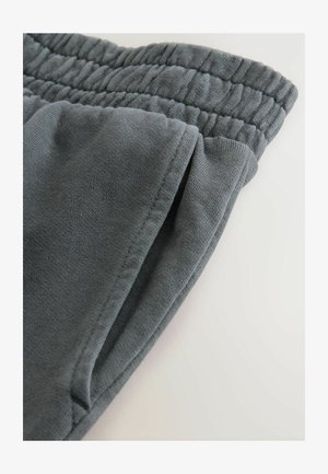 Prim-plan al unei buzunare de pantaloni de trening gri și al unui talie elastică pe un fundal deschis.