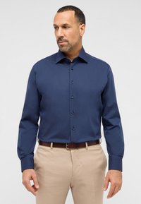 Camicia a maniche lunghe blu navy con chiusura a bottoni e colletto. Presenta un tessuto testurizzato e bottoni scuri a contrasto, abbinata a pantaloni beige.
