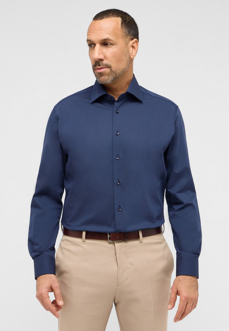 Camicia a maniche lunghe blu navy con chiusura a bottoni e colletto. Presenta un tessuto testurizzato e bottoni scuri a contrasto, abbinata a pantaloni beige.