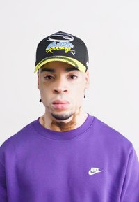 Svart caps med gule detaljer, med et design som leser "Powered Racing." Lilla sweatshirt med en diskret hvit Nike-logo.