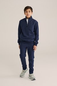 Mörkblå zip-up-tröja och matchande joggers i mjukt tyg med ribbade mudd- och sömnadselement. Utrustad med en kvartslång dragkedja.