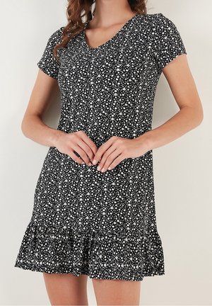 Femme portant une robe noire à manches courtes avec un motif floral blanc et un ourlet à volants, mains jointes à la taille, fond neutre.