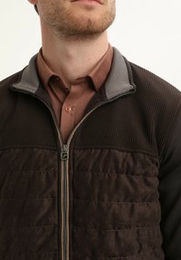Bruine ritsvest met een getextureerde geribde kraag en een effen gewatteerde voorkant. Is gemaakt van bruin en grijs stof, met een metalen rits en een_button-down shirt eronder.