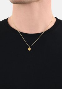 Collana con pendente a forma di cuore in oro su una delicata catena, indossata sopra una maglietta nera a girocollo. Il pendente è liscio e lucido, aggiungendo un tocco di eleganza.