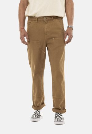 Homme portant un pantalon marron retroussé, une chemise blanche, des chaussures à enfiler à motifs à carreaux, une montre argentée et des bagues, debout devant un fond clair.