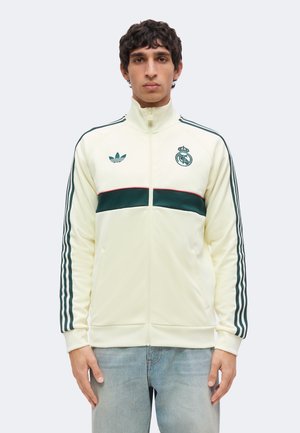 Joven con chaqueta deportiva color crema con rayas verdes, logotipos de Adidas y Real Madrid, y vaqueros azul claro, de pie frente a un fondo liso.