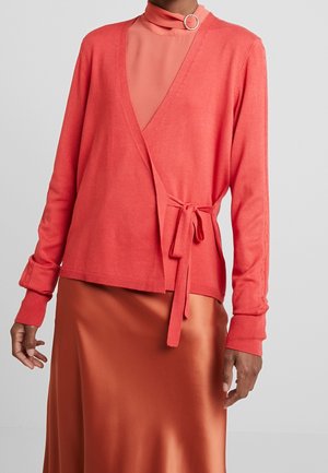 Cardigan en maille douce couleur corail avec des manches longues et un détail noué à la taille, associé à une jupe lisse en satin de couleur rouille.