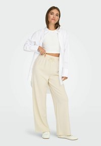 Žena v bílém krátkém topu bez rukávů, oversized bílém tričku, béžových širokých kalhotách a bílých teniskách, stojící s rukou na bocích.