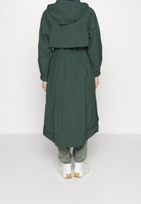 Dlouhá, tmavě zelená deštník s úzkým pasem, kapucí a šikmým lemem, doplněná světle zelenými kalhotami a bílou obuví.