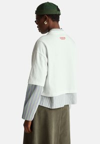 Biały krótki t-shirt z krótkim rękawem, z czerwonym logo "Stranger Things" na plecach, noszony na paskowanej koszulce z długim rękawem.