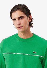Grøn sweatshirt med rund halsudskæring, kontrasterende hvid horisontal stribe og Lacoste-logo på brystet. Glat tekstur, casual pasform.