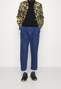 Camicia a fantasia verde e marrone, T-shirt nera, pantaloni jeans blu larghi e sneakers neri con accenti verdi.