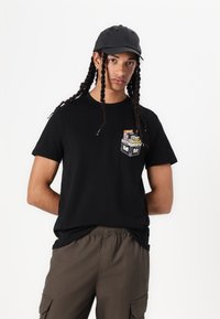 Black cotton t-shirt with a graphic pocket design, round neckline, and short sleeves, paired with gray cargo pants. Black cap on head.