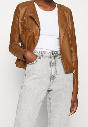 Personne portant une veste en cuir marron par-dessus un t-shirt blanc, avec un jean taille haute gris clair, une main dans la poche.