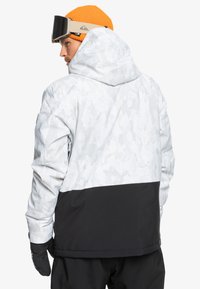 Schnee- und snowboardjacke in Schwarz-Weiß mit strukturiertem Muster, ausgestattet mit einer Kapuze, elastischen Bündchen und einem schwarzen unteren Abschnitt. Schwarze Handschuhe werden getragen.