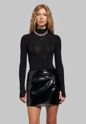 Jeune femme aux longs cheveux blonds portant un haut noir à manches longues, un collier chaîne argenté et une mini-jupe en cuir noir brillante sur fond gris.