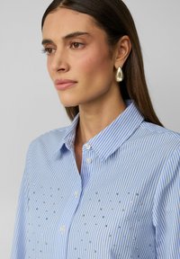 Blau-weiß gestreifte Bluse mit Kragen, ausgestattet mit silbernen Verzierungen an der Vorderseite. Glattes Material mit klassischem Knopfverschluss.