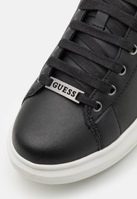 Svart lädersneaker med rund tå, svarta snörningar, silverfärgad loggtagg med texten "GUESS" och en vit gummisula. Texturerade ovandelar synliga.