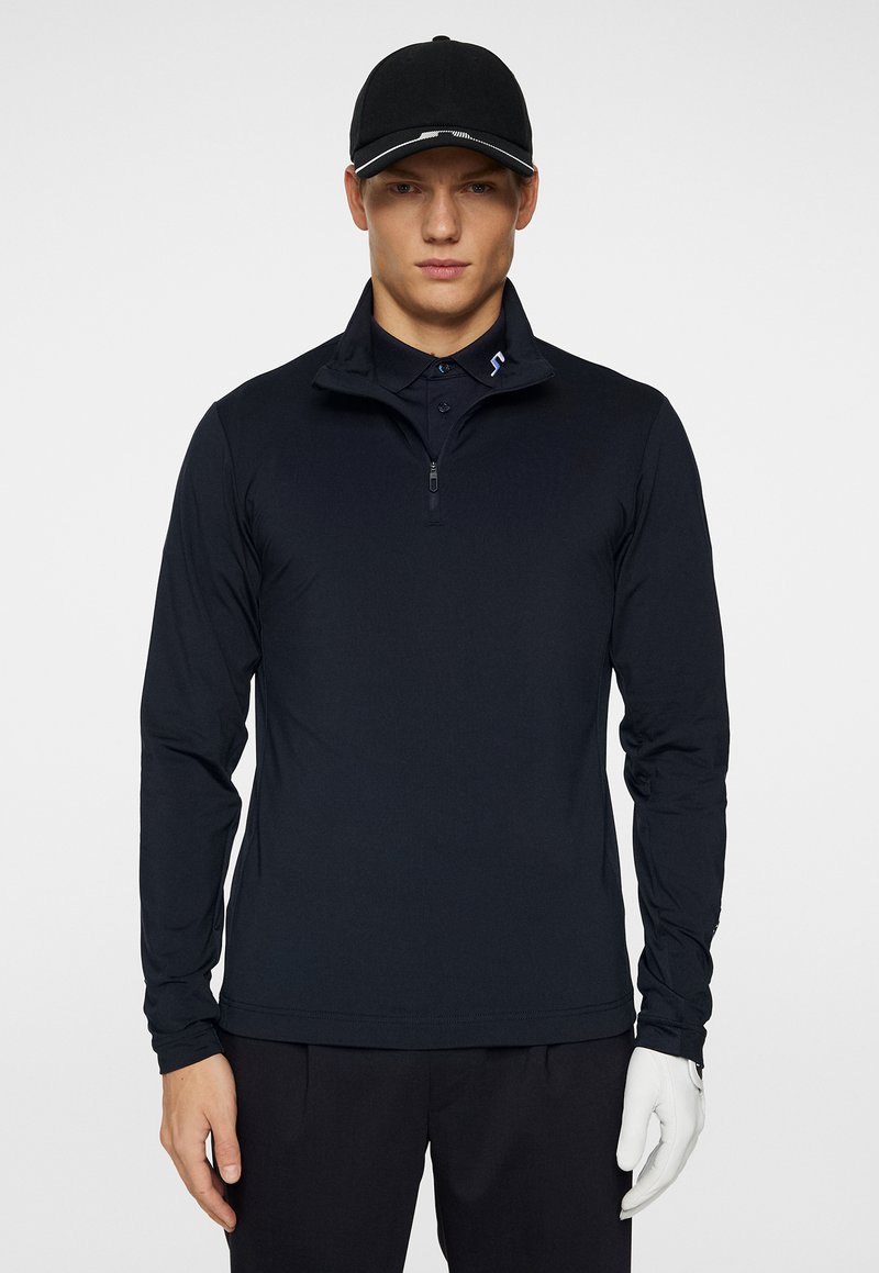 Polo a maniche lunghe blu navy con colletto a mezza zip, texture liscia e un sottile logo accentuato sul petto; indossato con pantaloni neri e un cappellino.