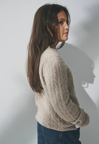 Beżowy sweter w splot kablowy wykonany z miękkiego materiału, z okrągłym dekoltem i długimi rękawami. Połączony z niebieskimi dżinsami.