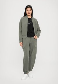 Vrouw staat in een olijfgroene zip-up jacket en cargobroek met een zwart T-shirt en witte sneakers tegen een effen lichte achtergrond.