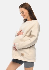 Beige Sweatshirt mit lässigem Schnitt, gerippten Bündchen und einem Lederlabel. Weiches Material, das für Komfort während der Schwangerschaft konzipiert ist.
