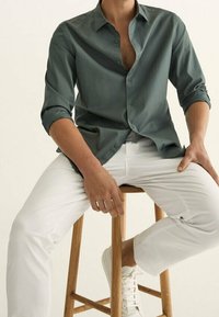 Groene button-up overhemd met lange mouwen, zittend op een houten kruk. Gecombineerd met witte broek en witte schoenen. Glad textuur van de stof.