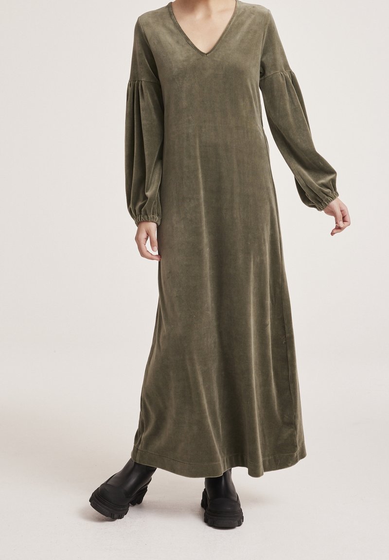 Robe maxi en velours vert olive avec un décolleté en V, des manches longues bouffantes et des poignets élastiques. Portée avec des bottines noires épaisses.