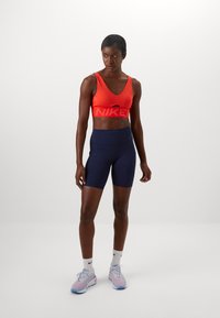 Red sports bra with a cut-out and large Nike logo, paired with navy high-waisted bike shorts. Light-colored athletic shoes and socks.